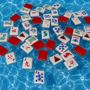 May include: A collection of white rectangular Mahjong tiles scattered across a blue water surface. The tiles feature various red, green, and blue symbols, numbers, and characters, including a tile labeled Joker with a blue figure illustration.