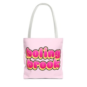 Pode incluir: Uma sacola rosa com o texto "boling brook" em uma fonte de bolhas com detalhes amarelos e rosa. A sacola tem alças brancas e provavelmente é feita de tecido.