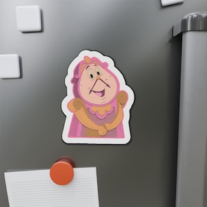 May include: A pink and tan cartoon clock character magnet. The clock has a smiling face, a pink hat, and a tan body. The magnet is attached to a gray surface with other magnets.
