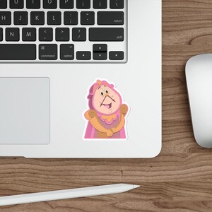 May include: A sticker featuring a cartoon character clock with a pink and gold color scheme. The clock has a smiling face and is wearing a pink cape. The sticker is on a laptop.