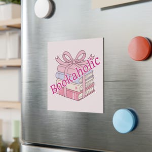 May include: A square magnet featuring a stack of three books tied with a pink ribbon bow. The books are colored in shades of pink, blue, and cream. The text Bookaholic is printed in a bold pink font diagonally across the center of the illustration.