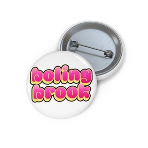 Pode incluir: Dois distintivos redondos brancos. Um mostra o texto "boling brook" em letras rosa bolha com contorno amarelo e borda preta. Os distintivos são redondos e têm um fecho de alfinete de metal.
