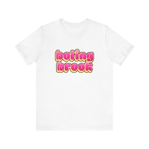 Pode incluir: Camiseta branca com o texto "boling brook" em fonte de bolhas. O texto é rosa com contorno amarelo. A camiseta é feita de um material macio e tem mangas curtas.