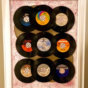 May include: Framed artwork featuring nine vintage vinyl records arranged in a grid. The records have colorful labels with various designs and text. The frame is white, and the background has a pink and white floral pattern.