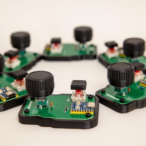 May include: A circular arrangement of five green printed circuit boards, each featuring a large black rotary knob, a square black push button, and a small blue electronic module with a USB port, all mounted on black plastic bases.