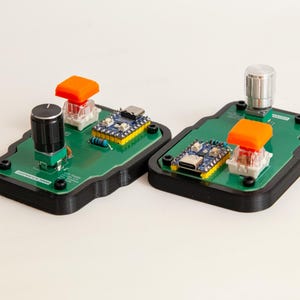 May include: Two electronic control modules featuring green circuit boards mounted on black bases. Each unit includes a blue microcontroller with a USB-C port, a bright orange square mechanical switch, and a silver rotary knob for manual input control.