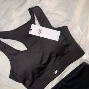 May include: Black sports bra and leggings set. The sports bra features a racerback design and the brand name "alo" printed in white on the band. A tag with the brand name is attached to the bra.