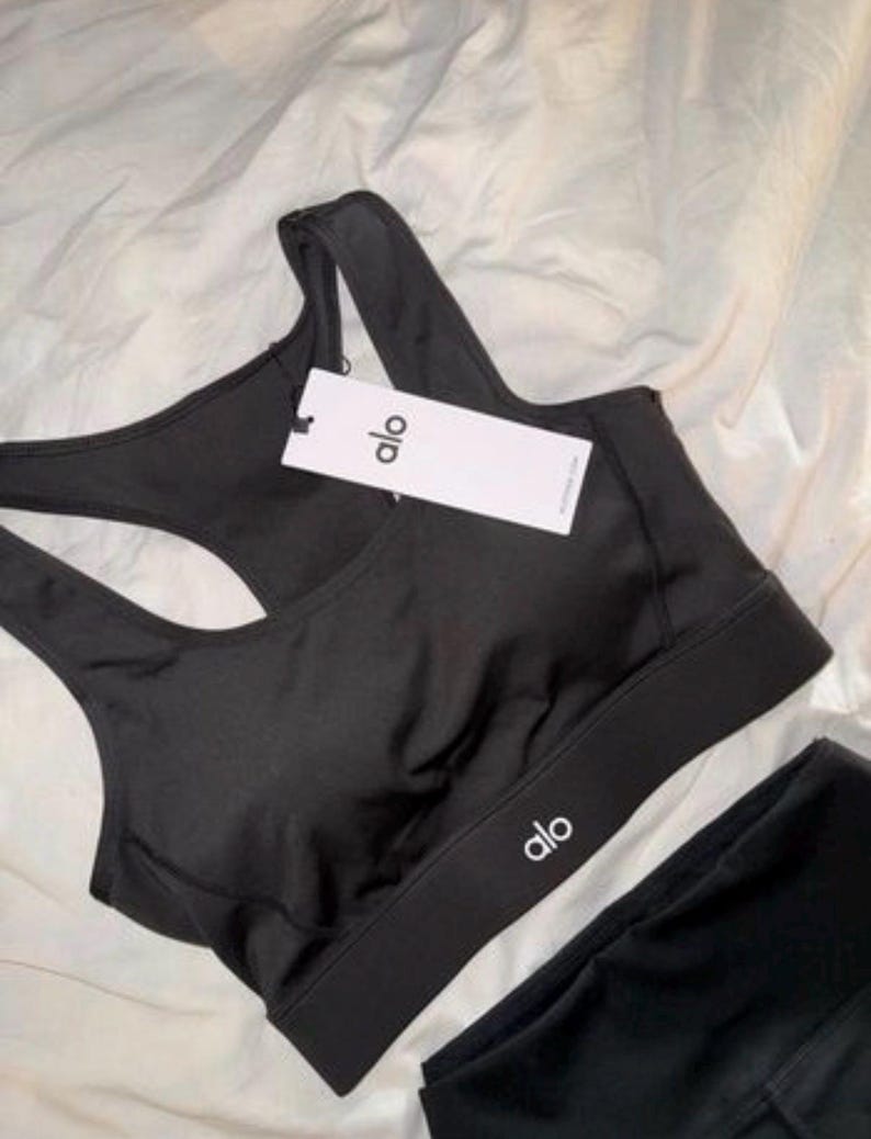 May include: Black sports bra and leggings set. The sports bra features a racerback design and the brand name "alo" printed in white on the band. A tag with the brand name is attached to the bra.