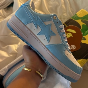 Peut inclure: Une basket bleue et blanche avec un motif d'étoile et d'éclair. La chaussure a une semelle rose clair et des lacets blancs. La chaussure est dans une boîte avec un motif camouflage.