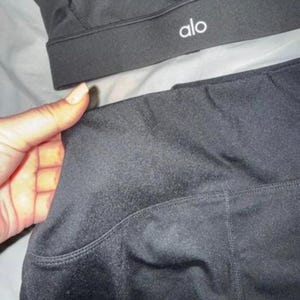 May include: Black sports bra and leggings set. The sports bra features the word "alo" in white. The leggings have a side pocket. The set is made from a smooth, matte fabric.