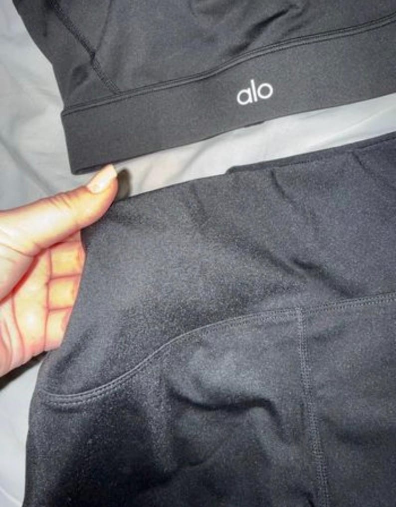 May include: Black sports bra and leggings set. The sports bra features the word "alo" in white. The leggings have a side pocket. The set is made from a smooth, matte fabric.
