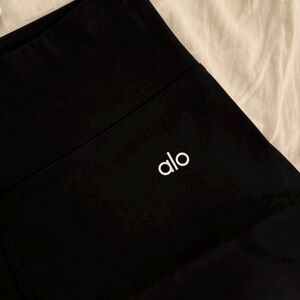 May include: Close-up of black athletic leggings with a high waistband. The white brand name "alo" is printed on the left leg. The leggings are made of a smooth, opaque fabric.