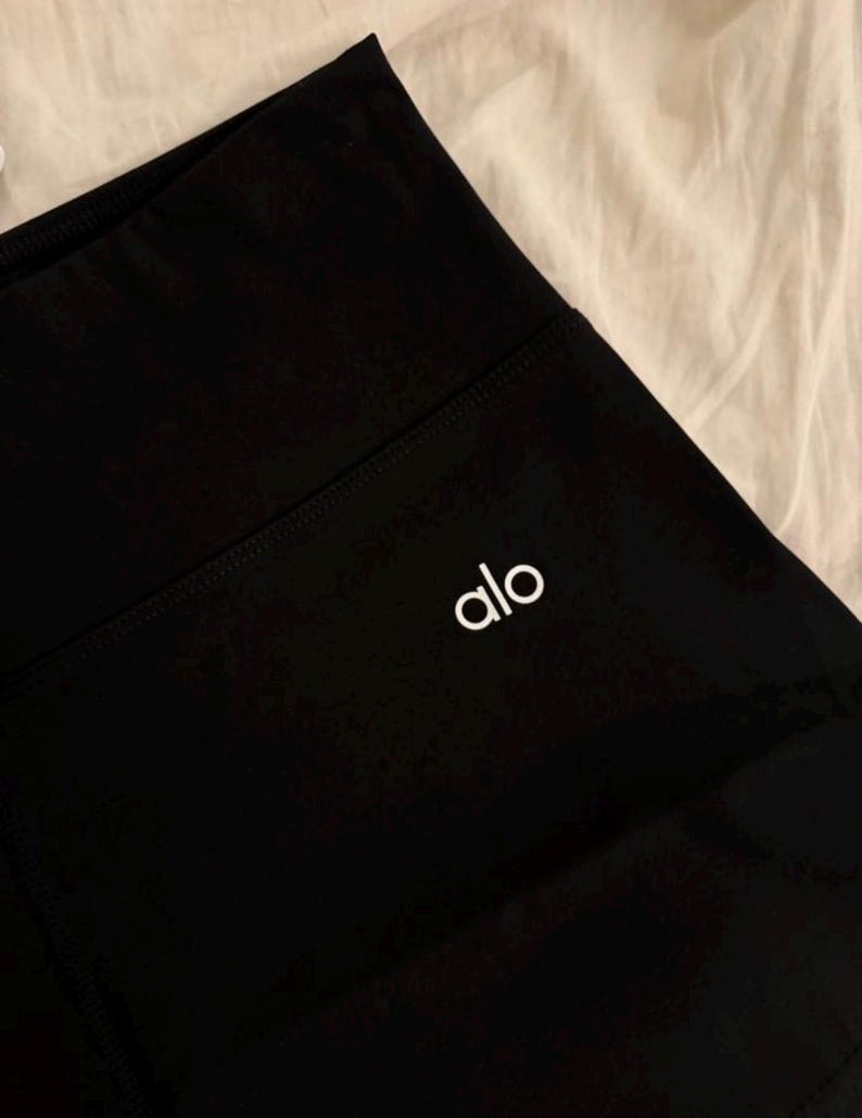 May include: Close-up of black athletic leggings with a high waistband. The white brand name "alo" is printed on the left leg. The leggings are made of a smooth, opaque fabric.