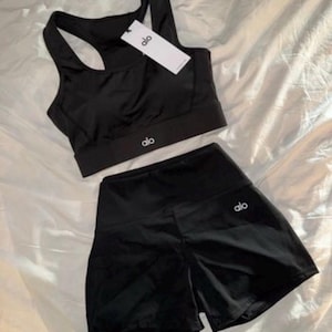 May include: Black athletic set comprising a sports bra and shorts. The sports bra has a racerback design and the shorts are high-waisted. Both items display the brand logo. The set is made of a shiny, smooth material.