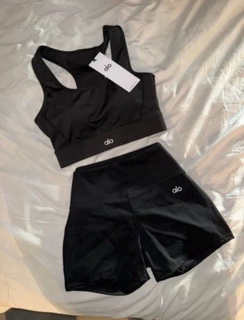 May include: Black athletic set comprising a sports bra and shorts. The sports bra has a racerback design and the shorts are high-waisted. Both items display the brand logo. The set is made of a shiny, smooth material.