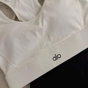 May include: White sports bra with a racerback design and the brand name "alo" in black lettering. The bra is paired with black athletic leggings. The image is well-lit, showcasing the clean lines and minimalist aesthetic of the activewear.