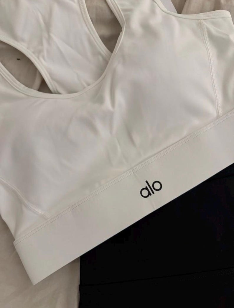 May include: White sports bra with a racerback design and the brand name "alo" in black lettering. The bra is paired with black athletic leggings. The image is well-lit, showcasing the clean lines and minimalist aesthetic of the activewear.