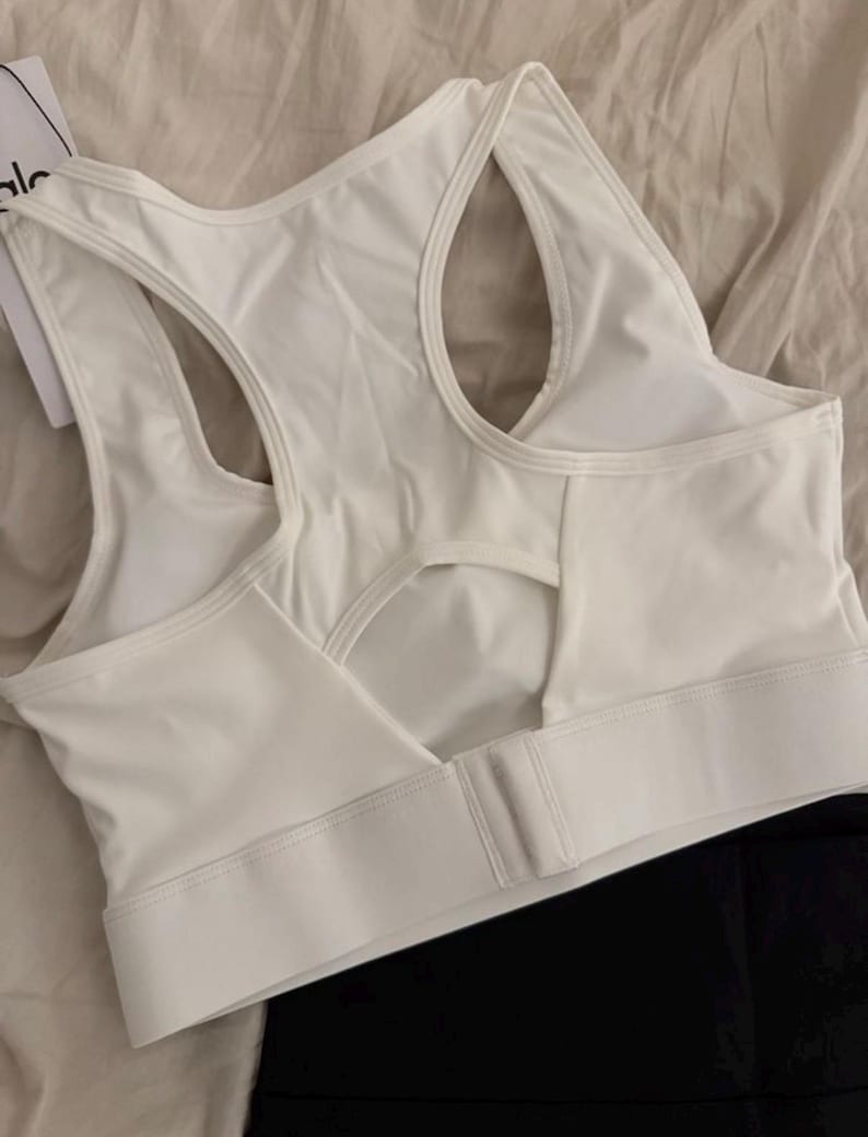 May include: White sports bra with a racerback design and a keyhole cutout in the back. The bra has a wide band under the bust and a hook-and-eye closure. The brand name "alo" is visible on a tag.