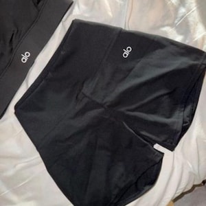 May include: Black athletic shorts and a sports bra, both featuring the brand name "alo" in white. The shorts are high-waisted, and the sports bra has a thick band. The items are made of a smooth, shiny material, suitable for exercise.