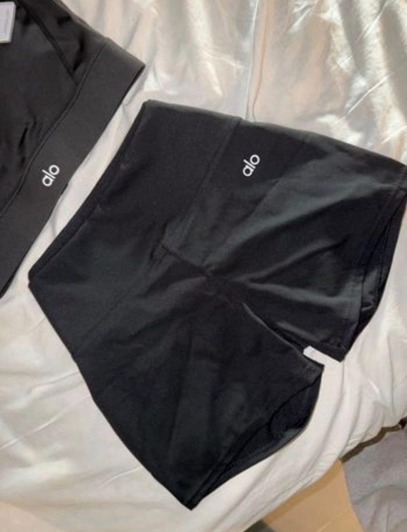 May include: Black athletic shorts and a sports bra, both featuring the brand name "alo" in white. The shorts are high-waisted, and the sports bra has a thick band. The items are made of a smooth, shiny material, suitable for exercise.