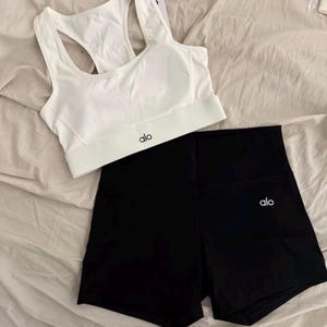 May include: A white sports bra and black athletic shorts set. The sports bra has a racerback design and the brand name "alo" printed in black. The shorts are high-waisted and also feature the "alo" logo in white.