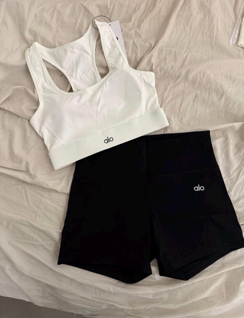 May include: A white sports bra and black athletic shorts set. The sports bra has a racerback design and the brand name "alo" printed in black. The shorts are high-waisted and also feature the "alo" logo in white.