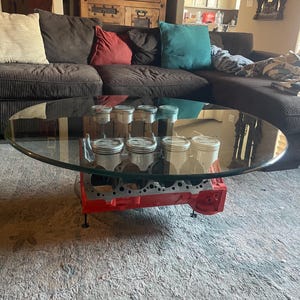 May include: Oval glass coffee table with a red engine block base. The base supports the glass top and features several pistons. The table is in a living room setting with a brown sectional sofa.