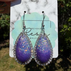 May include: A pair of teardrop-shaped earrings with a purple and glittery design. The earrings are framed with a silver-colored, rhinestone-studded border and have silver-colored hooks. The earrings are displayed on a white and gray marble-patterned card.