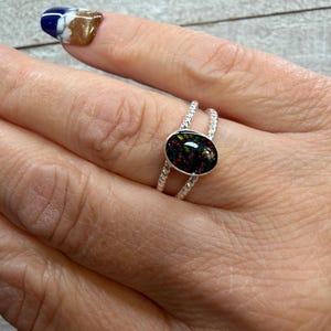 May include: A silver ring with a black opal stone. The oval-shaped opal has flecks of red, green, and gold. The ring band has a twisted rope design. The ring is on a finger with a painted nail.