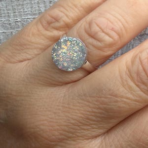 May include: A silver ring with a large, round, iridescent, glittery stone. The stone reflects multiple colors, including pink, green, and blue. The ring is shown on a person's finger.
