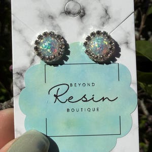 May include: A pair of iridescent stud earrings with a silver-colored, beaded border. The earrings are displayed on a white and gray marble-patterned card with the text "BEYOND Resin BOUTIQUE".