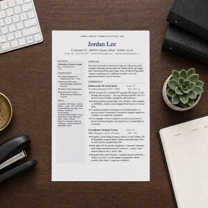May include: A resume titled "Jordan Lee" is centered on a dark wood desk, surrounded by office supplies. A white keyboard, stapler, gold paperclip dish, succulent plant, and a notebook are visible.