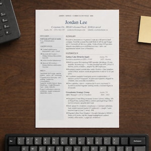 May include: A resume with the name "Jordan Lee" is displayed on a wooden desk, alongside a black telephone, a keyboard, a small brass dish with paper clips, and a yellow sticky note. The resume includes text.