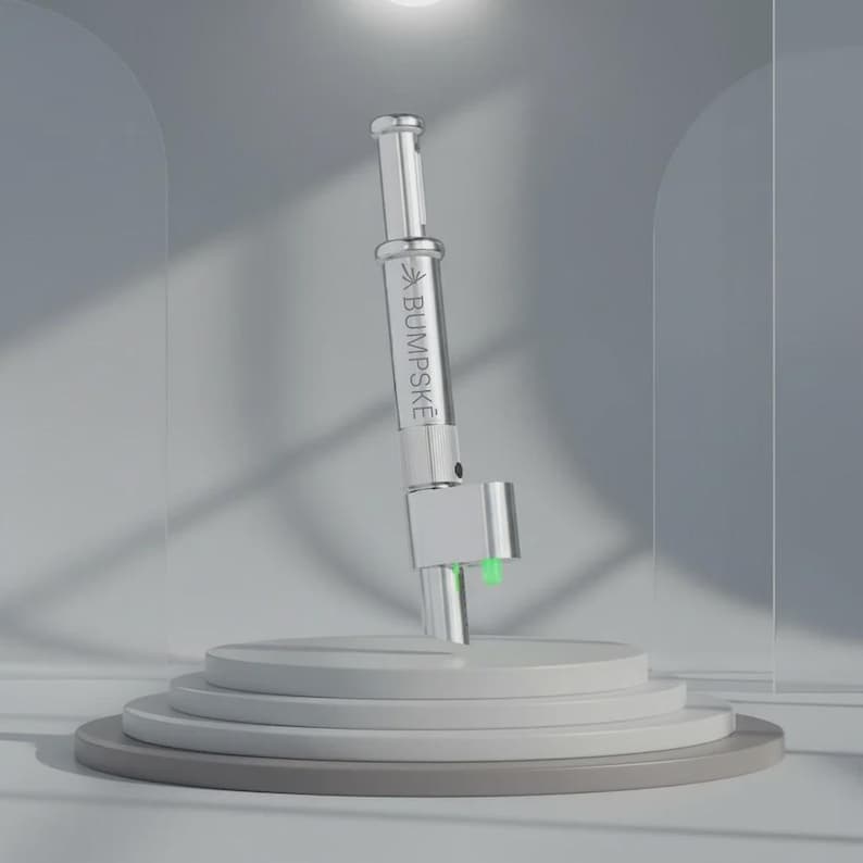 May include: A silver-toned cylindrical metal device with a clear glass section featuring the text BUMPSKE printed in black. The tool has a metallic base with two small, bright green protrusions extending from the bottom, set against a neutral background.
