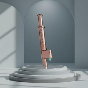 May include: A rose gold metal cylindrical tool with a plunger mechanism at the top and a small green tip at the base. The side of the device features the engraved text BUMPSKE with a small leaf icon above the lettering, presented in a vertical orientation.