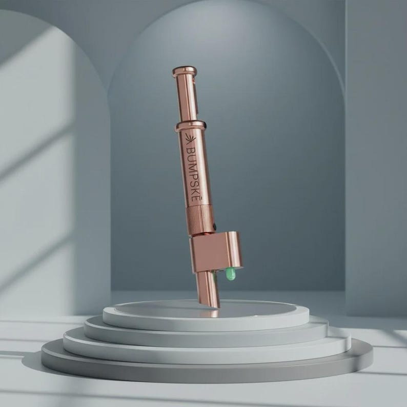 May include: A rose gold metal cylindrical tool with a plunger mechanism at the top and a small green tip at the base. The side of the device features the engraved text BUMPSKE with a small leaf icon above the lettering, presented in a vertical orientation.