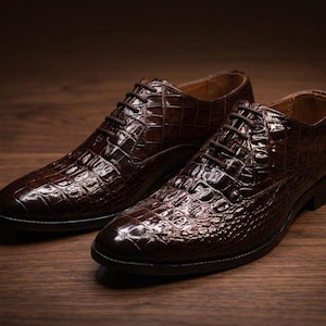 Può includere: Un paio di scarpe eleganti marrone scuro caratterizzate da una texture lucida a motivo pelle di coccodrillo. Queste scarpe stringate presentano una punta arrotondata, un tacco basso e lacci marrone scuro che richiamano la finitura lucida.