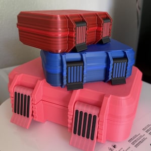 May include: A stack of three rectangular storage cases in red, blue, and pink. Each case features a textured, ribbed surface, rounded corners, and two black vertical latch mechanisms on the front for secure closure and storage organization.