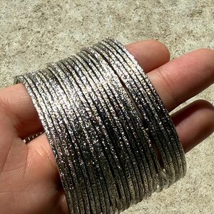 May include: A stack of silver-colored bangles with a textured surface. The bangles are thin and appear to be made of metal. The bracelets are stacked together, creating a layered effect.