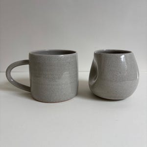 May include: Two ceramic mugs with a speckled light gray finish. The mug on the left features a standard cylindrical shape with a curved handle, while the mug on the right has a rounded, asymmetrical body with a distinct indentation on one side.