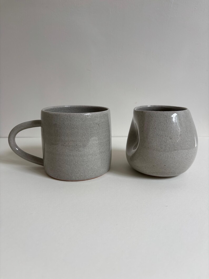 May include: Two ceramic mugs with a speckled light gray finish. The mug on the left features a standard cylindrical shape with a curved handle, while the mug on the right has a rounded, asymmetrical body with a distinct indentation on one side.