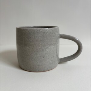 May include: A cylindrical ceramic mug in a light gray color with a visible crackle glaze finish and dark speckles throughout the surface. The mug features a smooth, curved handle on the right side and a flat base with a thin terracotta-colored rim.