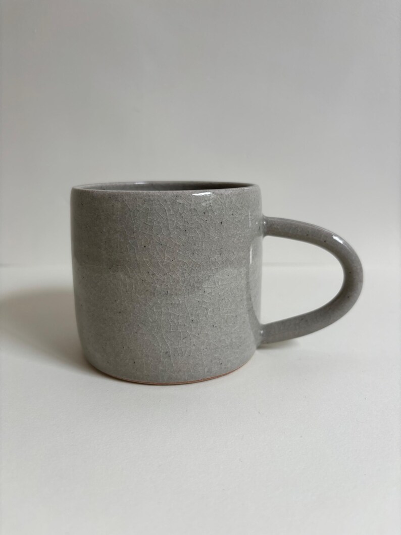 May include: A cylindrical ceramic mug in a light gray color with a visible crackle glaze finish and dark speckles throughout the surface. The mug features a smooth, curved handle on the right side and a flat base with a thin terracotta-colored rim.