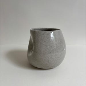May include: A light gray ceramic cup with a speckled texture and a visible crackle glaze finish. The cup features an asymmetrical, rounded shape with a distinct indentation on one side, creating a unique, organic form without a handle.