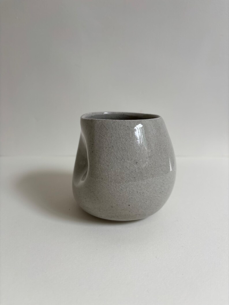 May include: A light gray ceramic cup with a speckled texture and a visible crackle glaze finish. The cup features an asymmetrical, rounded shape with a distinct indentation on one side, creating a unique, organic form without a handle.