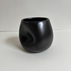 May include: A matte black ceramic cup with a rounded, bulbous shape and a distinct, singular indentation on one side. The vessel features a smooth, dark finish and a wide, open circular rim, presenting a solid, monochromatic appearance throughout.