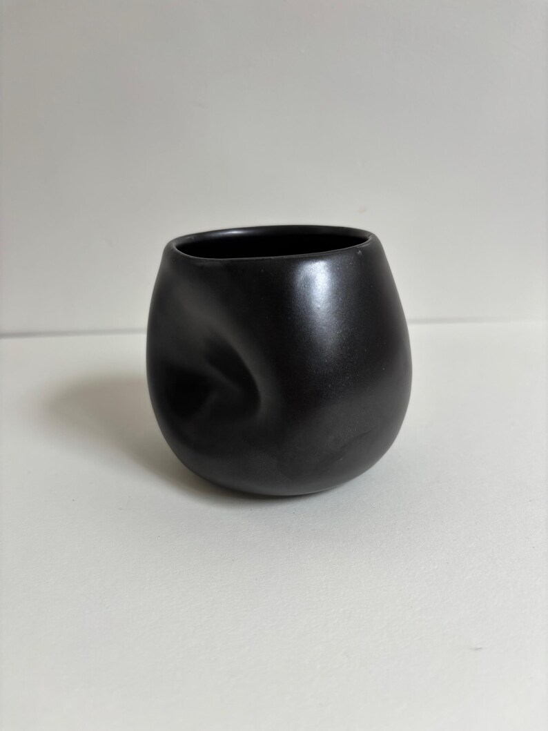May include: A matte black ceramic cup with a rounded, bulbous shape and a distinct, singular indentation on one side. The vessel features a smooth, dark finish and a wide, open circular rim, presenting a solid, monochromatic appearance throughout.