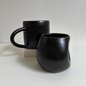 May include: A matte black ceramic mug with a rounded handle and a matching matte black ceramic cup featuring an irregular, organic, indented shape. Both items have a smooth, dark finish and are positioned together to show their distinct forms.