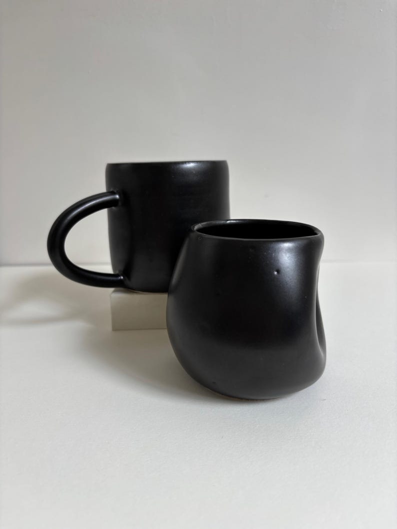 May include: A matte black ceramic mug with a rounded handle and a matching matte black ceramic cup featuring an irregular, organic, indented shape. Both items have a smooth, dark finish and are positioned together to show their distinct forms.