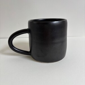 May include: A matte black ceramic mug with a smooth, cylindrical body and a curved handle on the left side. The mug features a solid, dark finish with subtle horizontal indentations along the exterior surface, creating a simple, uniform appearance.